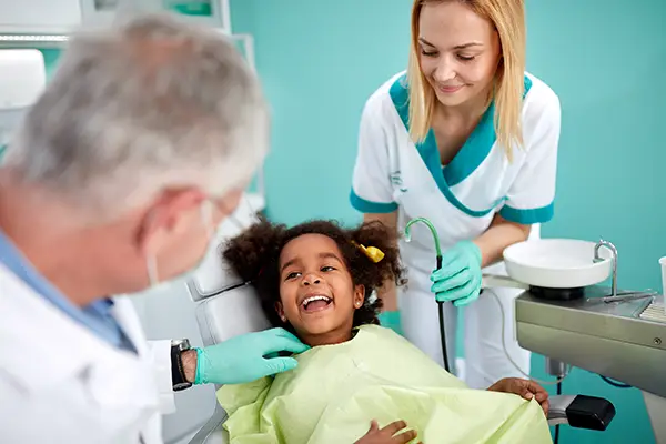 Adorable and young black girl sitting in a dental chair and smiling at her older white dentist and white dental assistant.