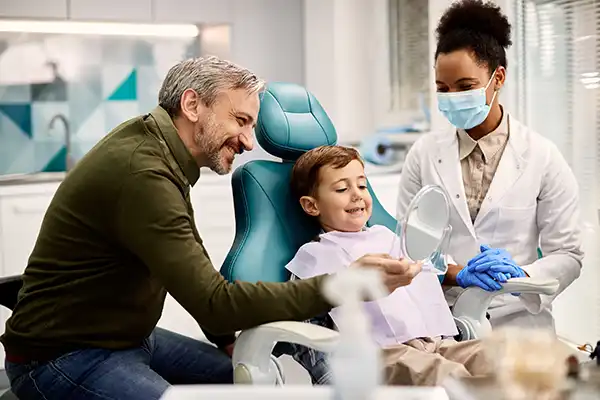 Father with facial hair holding a mirror for his happy, young son sitting in a dental chair while his female dentist wearing a mask happily watches, at McHenry Smile Center.